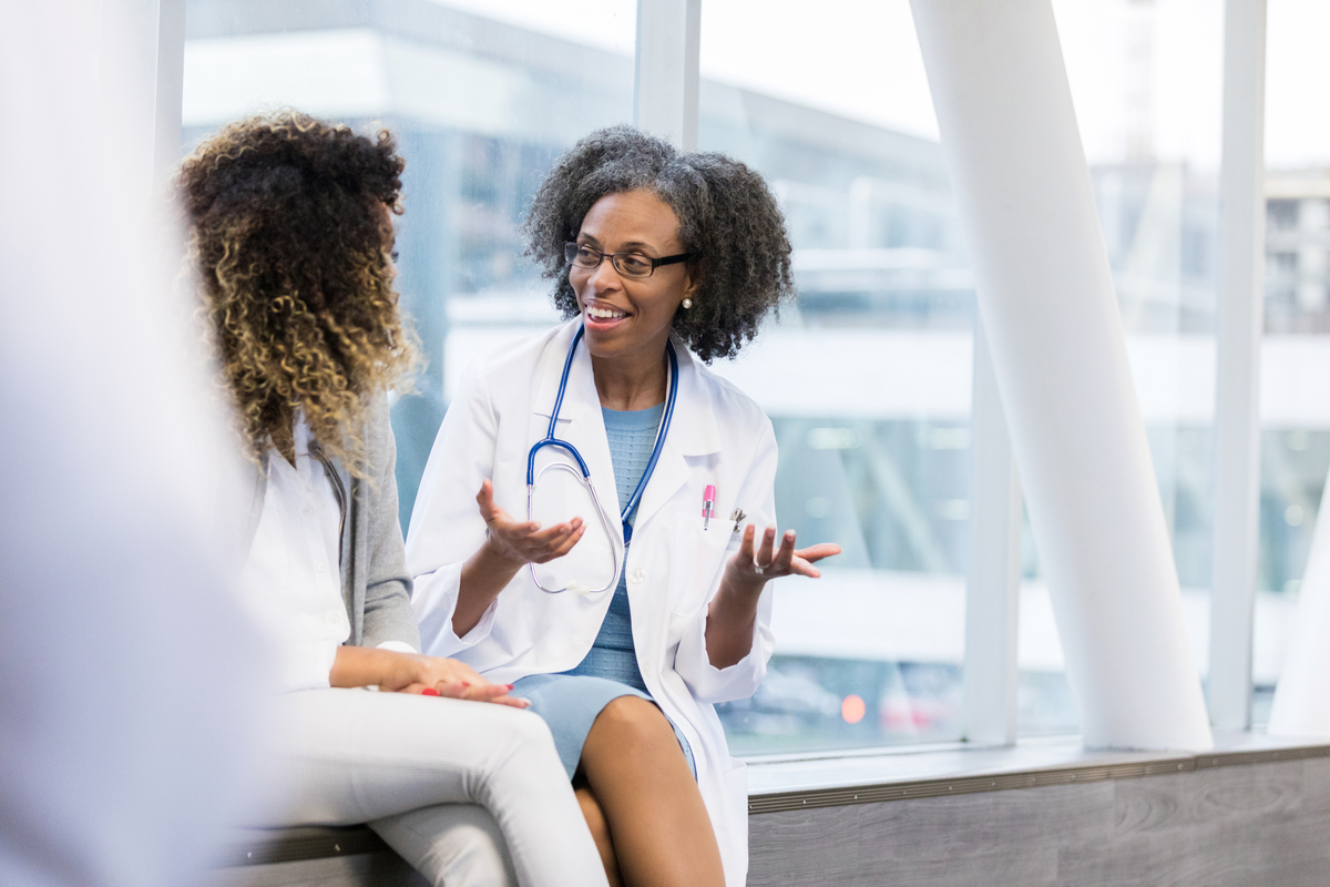 Mature female doctor gestures while talking with a young woman while in a hospital corridor.